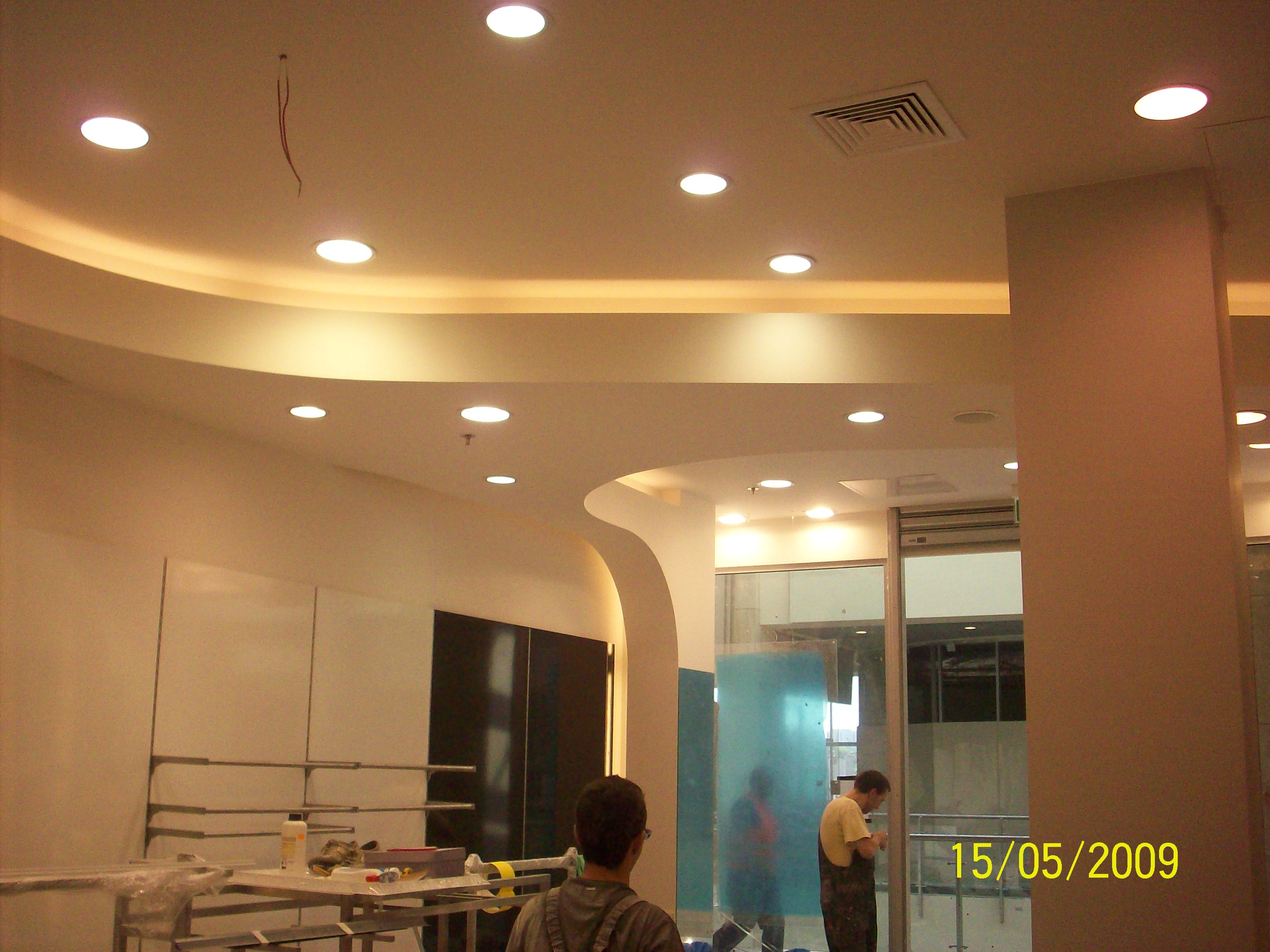 Wnętrze w trakcie wykańczania: widoczny sufit z oświetleniem punktowym i podświetleniem ledowym, fragment ściany z zamontowanymi półkami, oraz dwójka pracowników pracujących przy szklanej ściance...